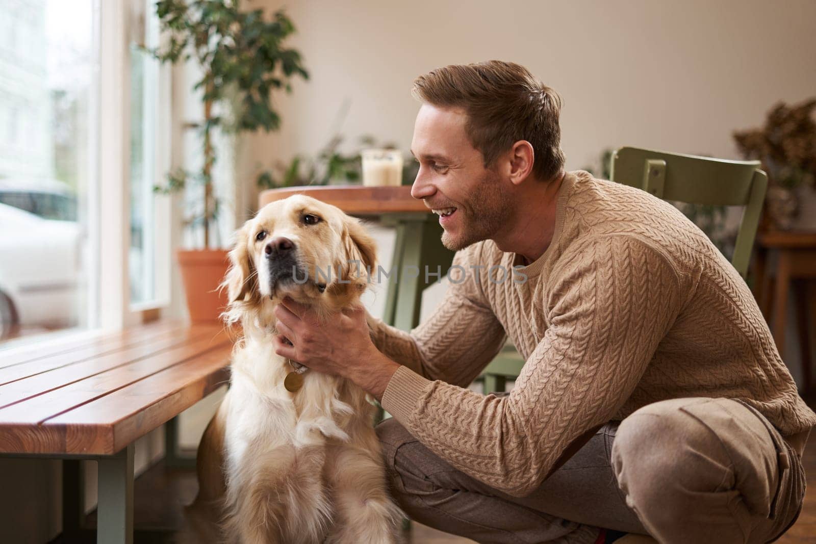 Portrait of happy smiling handsome man, pets his golden retriever and laughs. Young guy with his dog in a cafe by Benzoix