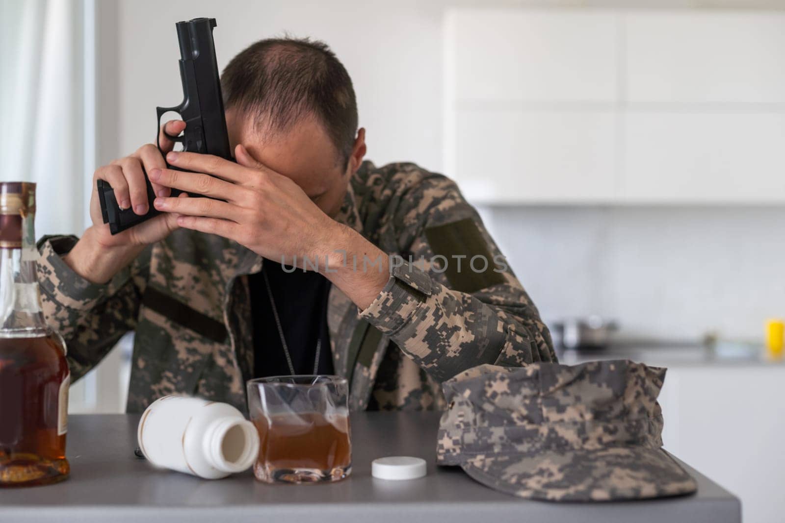 A man in camouflage suffers from depression after returning from the army. He uses alcohol and narcotic tablets. He is tormented by heavy memories. On the table is a gun. High quality photo