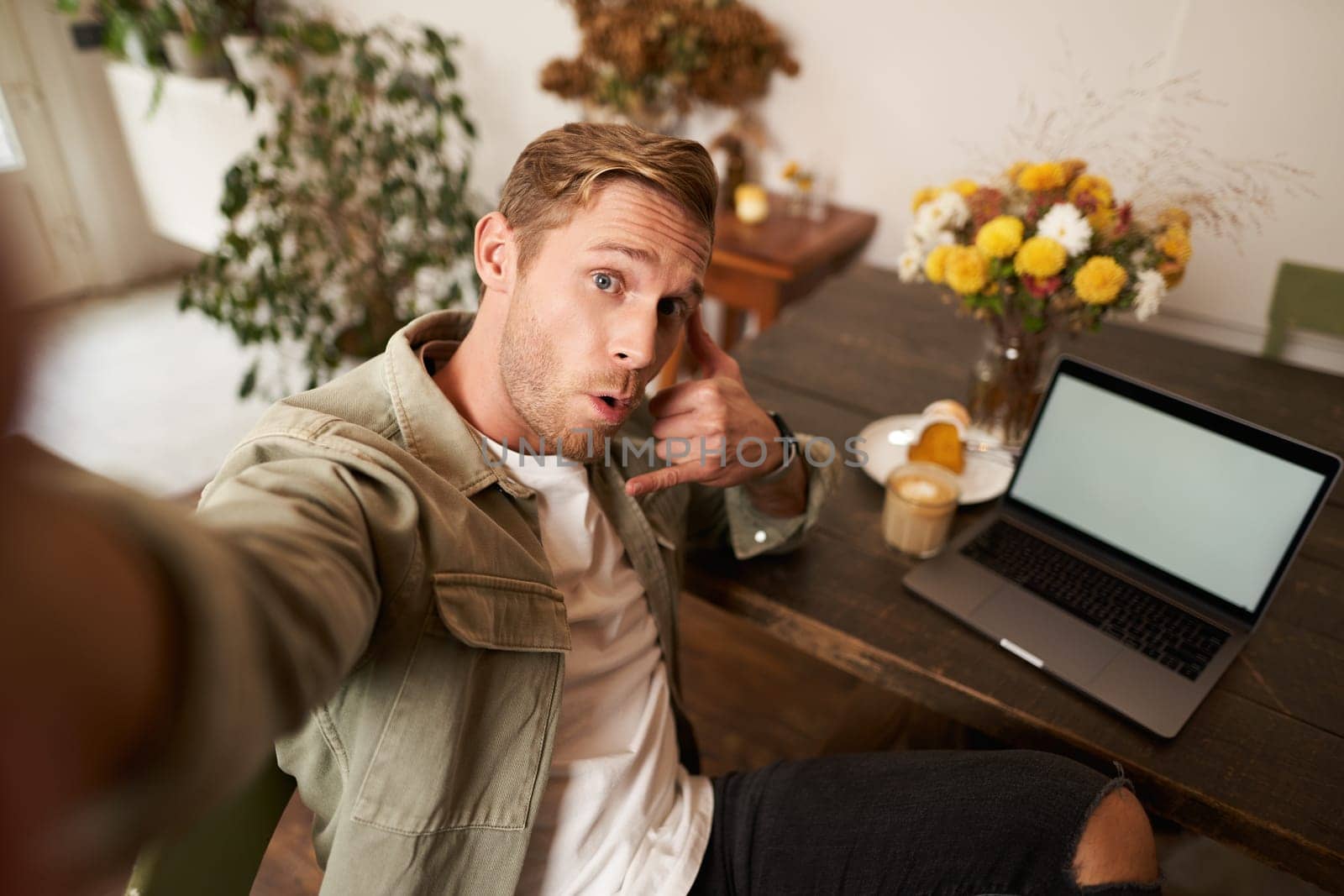 Handsome stylish young man takes selfie in cafe while working or studying remotely with laptop, shows call phone hand sign and smiling at mobile phone camera by Benzoix