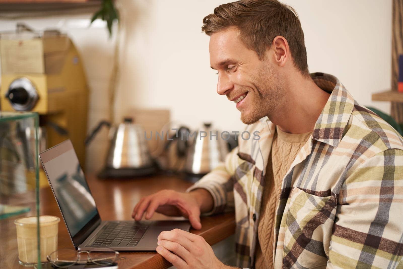 Portrait of handsome businessman, young digital nomad, sitting in cafe and looking at his laptop screen, working remotely, doing freelance project from coffee shop.