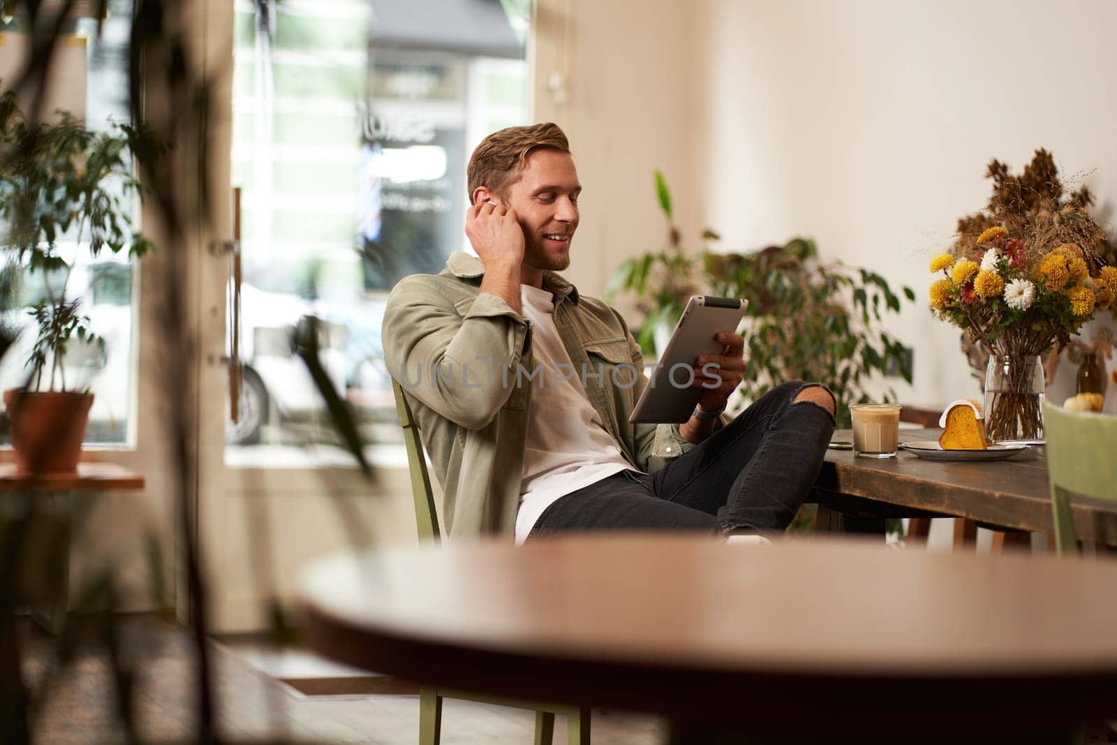 Portrait of handsome happy guy, young man sits in cafe, watching videos on digital tablet, wearing wireless headphones, laughing and smiling, spending time in coffee shop by Benzoix