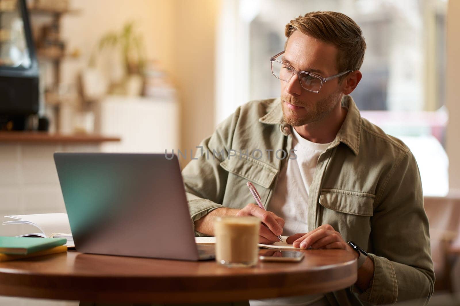 Portrait of handsome young guy in glasses, man studying, taking notes, looking at laptop screen during online lesson, attends a meeting and writing down information, sitting in cafe by Benzoix