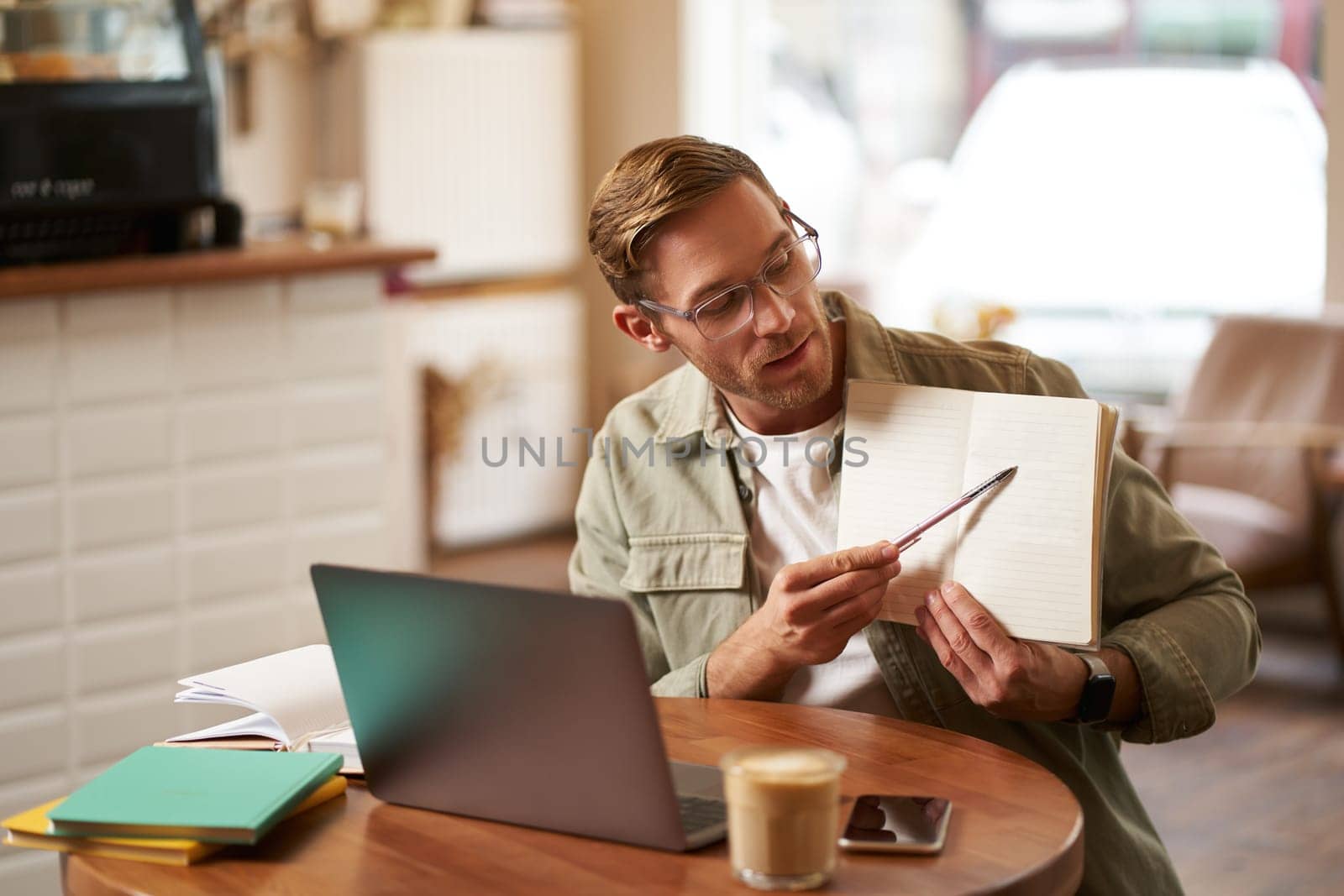 Portrait of handsome young man giving lessons online, showing exercises in his notebook, looking at laptop, recording video, sitting in cafe, quite co-working space by Benzoix