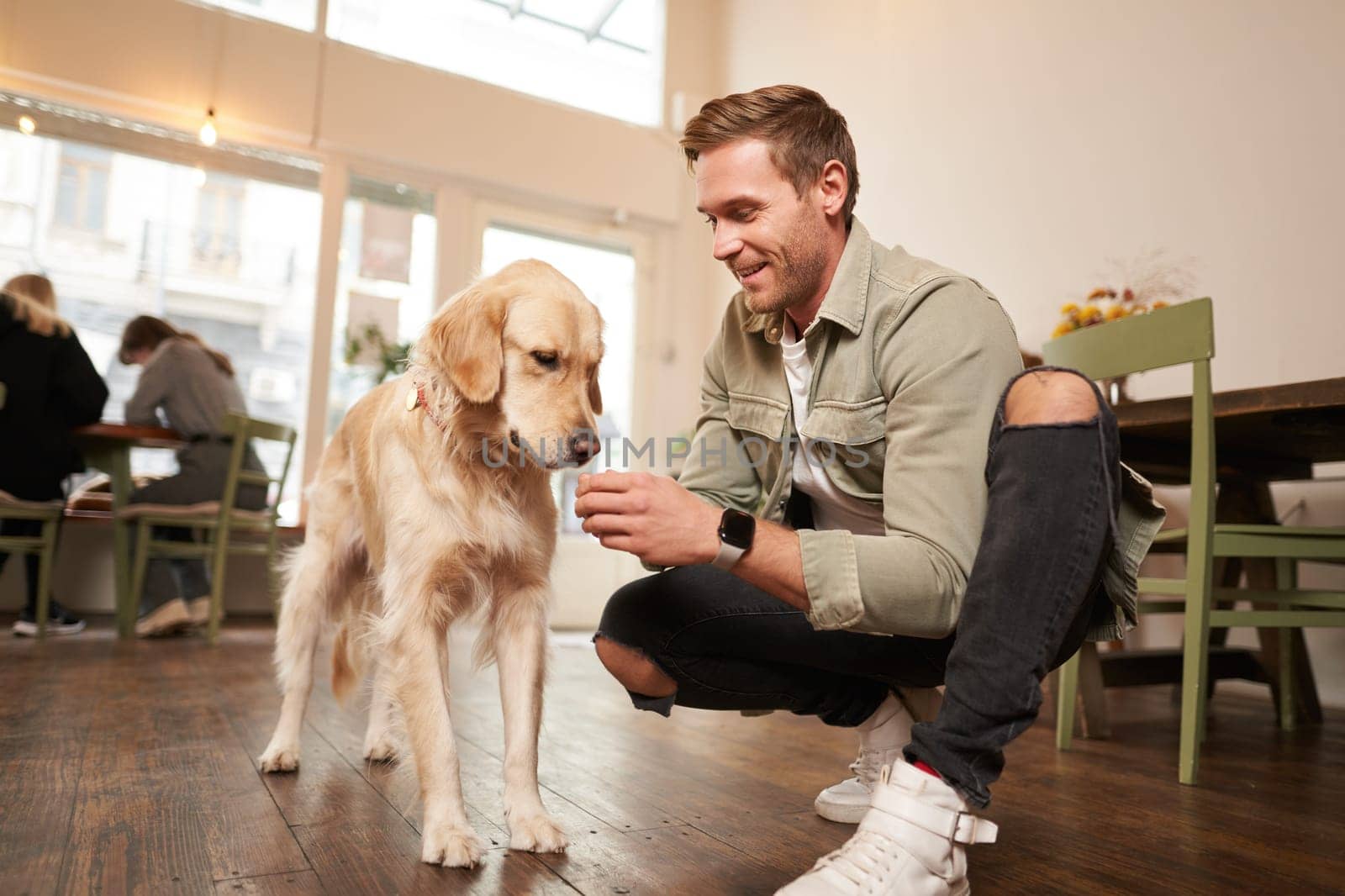 Portrait of smiling handsome man with his dog, sitting on floor in cafe with golden retriever, giving a treat.