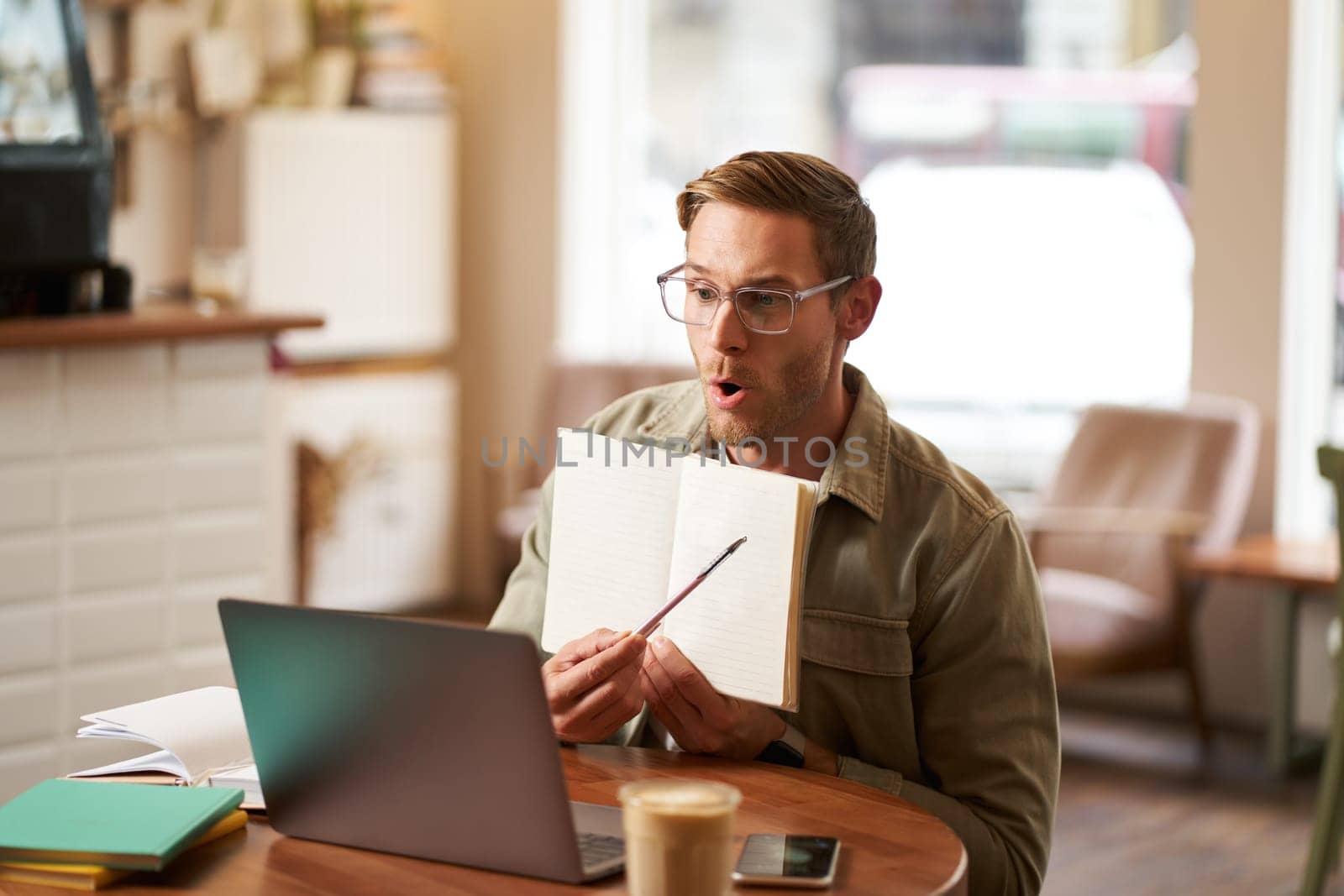 Portrait of handsome young man giving lessons online, showing exercises in his notebook, looking at laptop, recording video, sitting in cafe, quite co-working space.