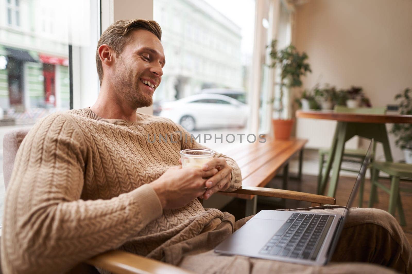 Portrait of handsome young man sits in cafe, drinks coffee and watches video on laptop, looking at screen with happy smile, relaxing in co-working space by Benzoix