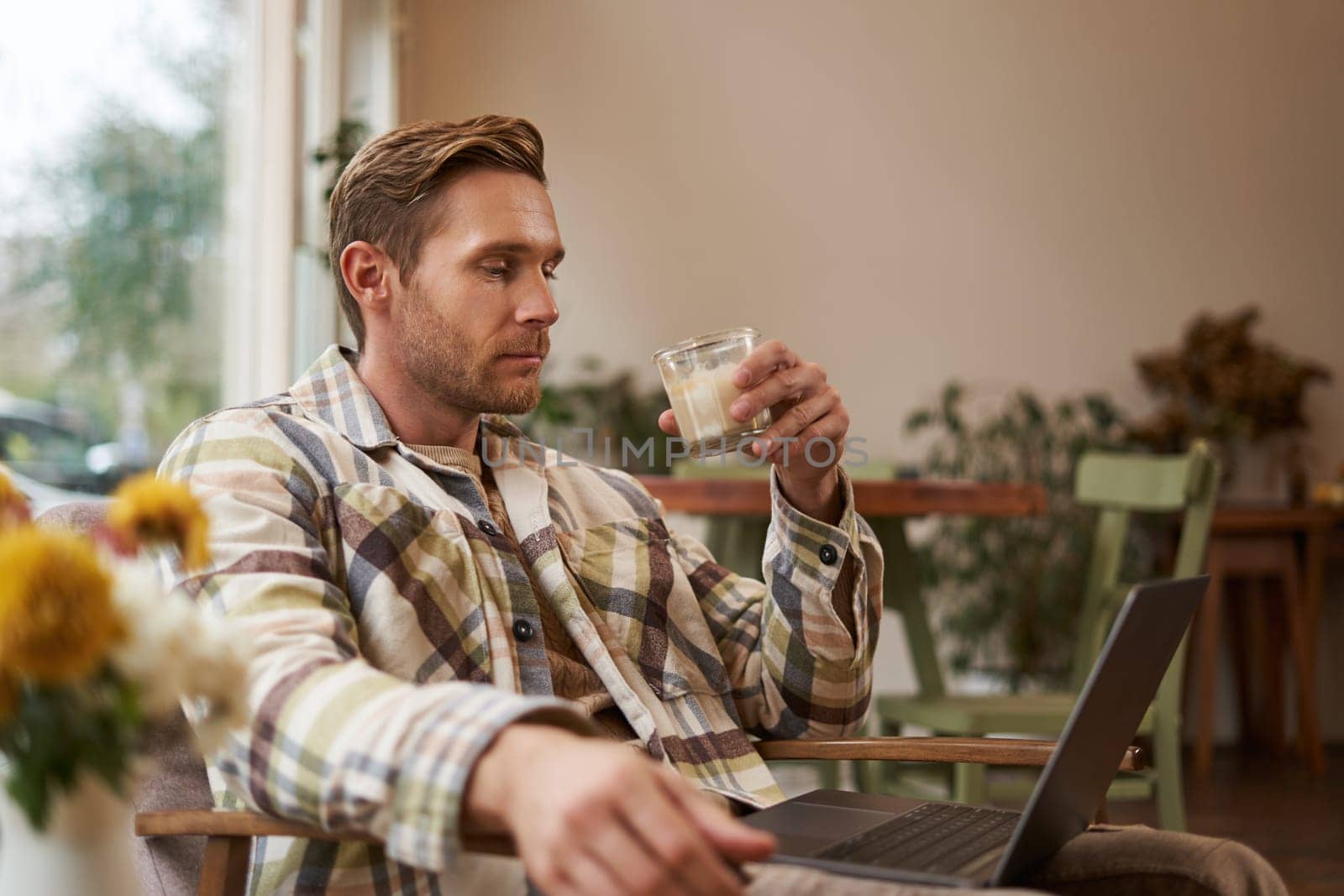 Portrait of young freelancer, a man in casual clothes, sitting in cafe and drinking coffee, working remotely on laptop, digital nomad doing his project outdoors in co-working space by Benzoix