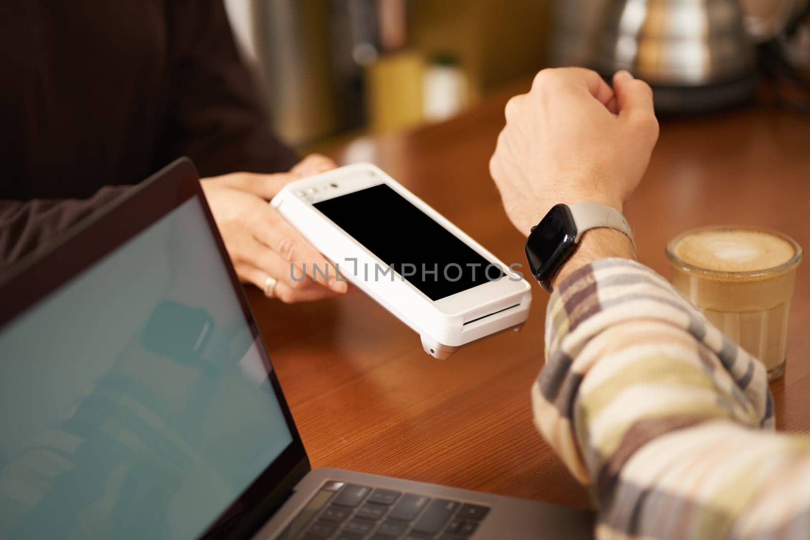 Close up shot of man paying with his watch, holding wrist near POS terminal for contactless payment, sitting in cafe co-working space with laptop by Benzoix