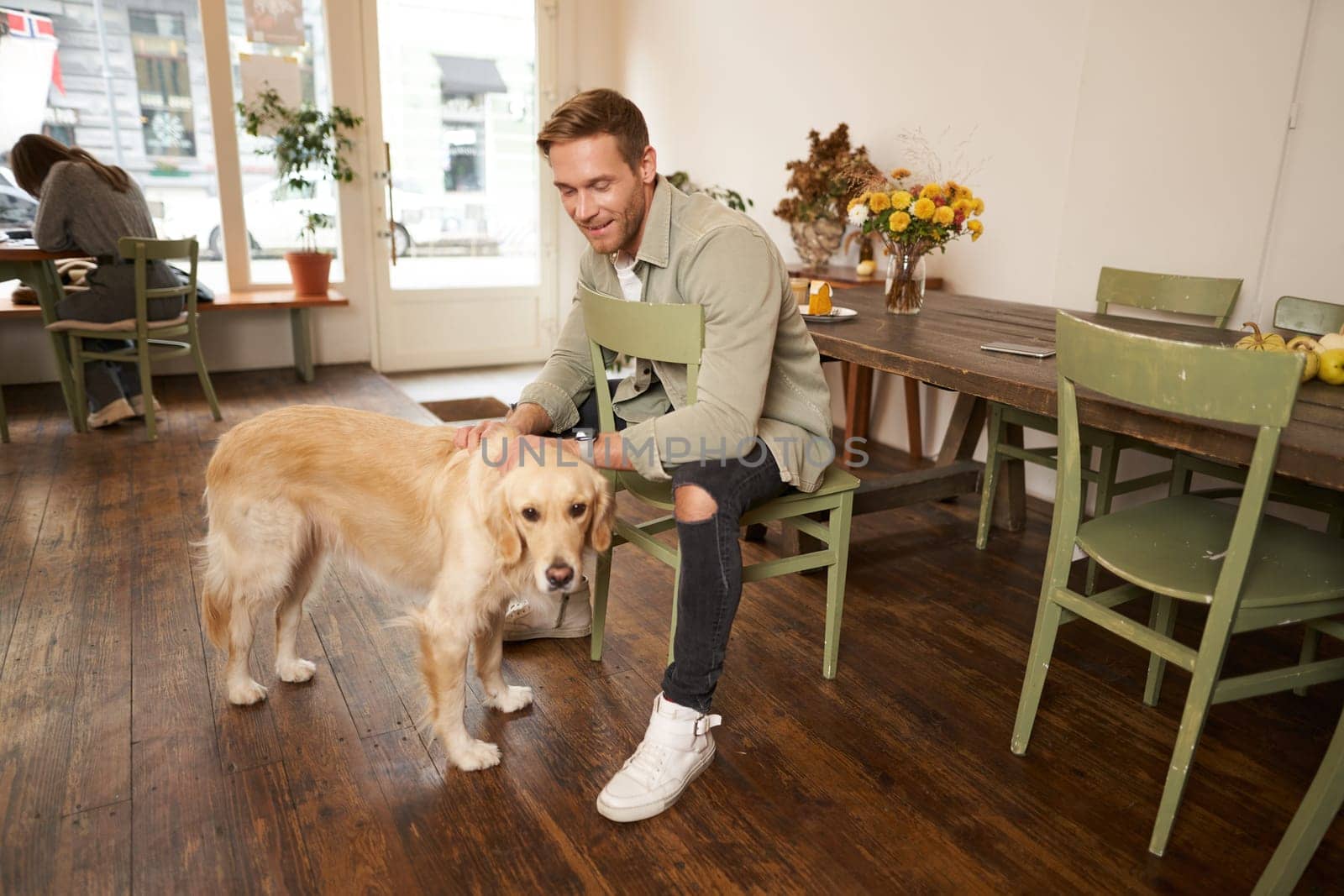 Lifestyle portrait of handsome smiling man sitting in pet friendly cafe with his beautiful dog, petting golden retriever, waiting for an order.