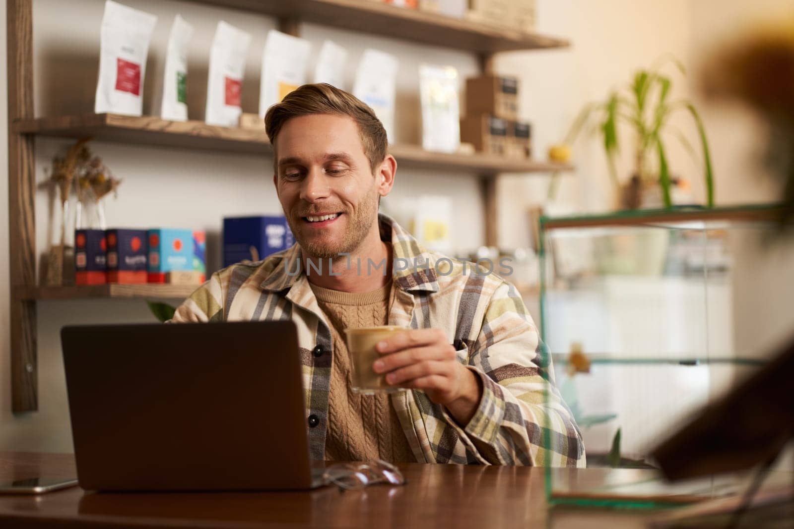 Lifestyle portrait of young businessman, freelancer sitting in cafe, drinking coffee and using laptop, working on online project, digital nomad doing his task, has remote job, looking satisfied.