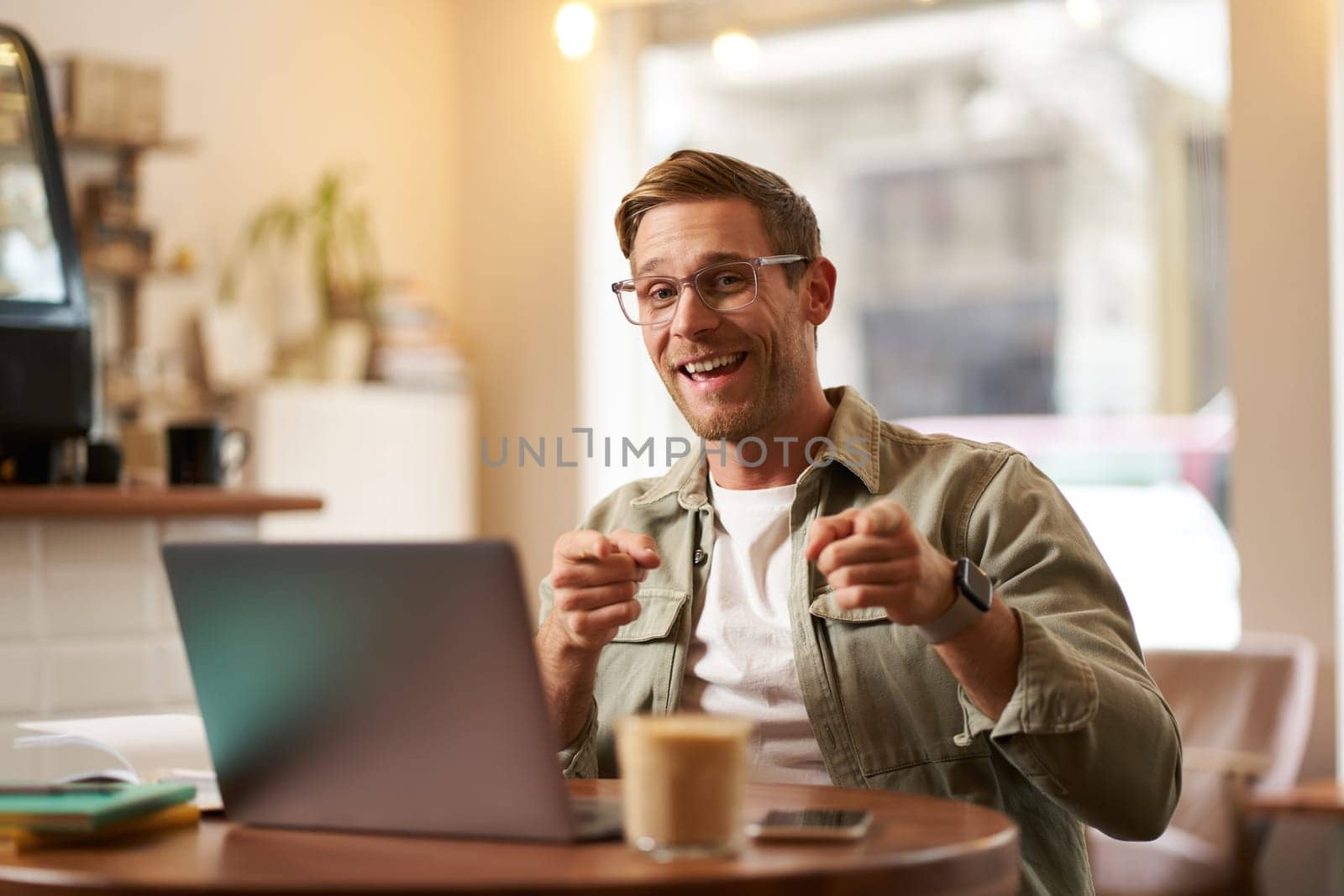 Portrait of cheerful guy in glasses, sits in cafe and works on laptop, pointing fingers at camera, inviting you, recommends you something, promoting product or public space.