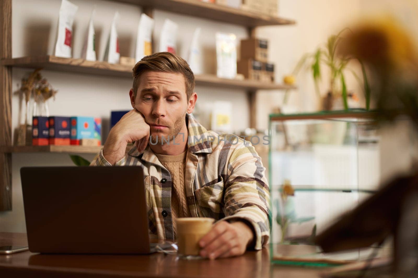 Portrait of man looking disappointed at laptop screen, working remotely, freelancer sitting in cafe, drinking coffee, reading message on monitor with unamused, upset face by Benzoix