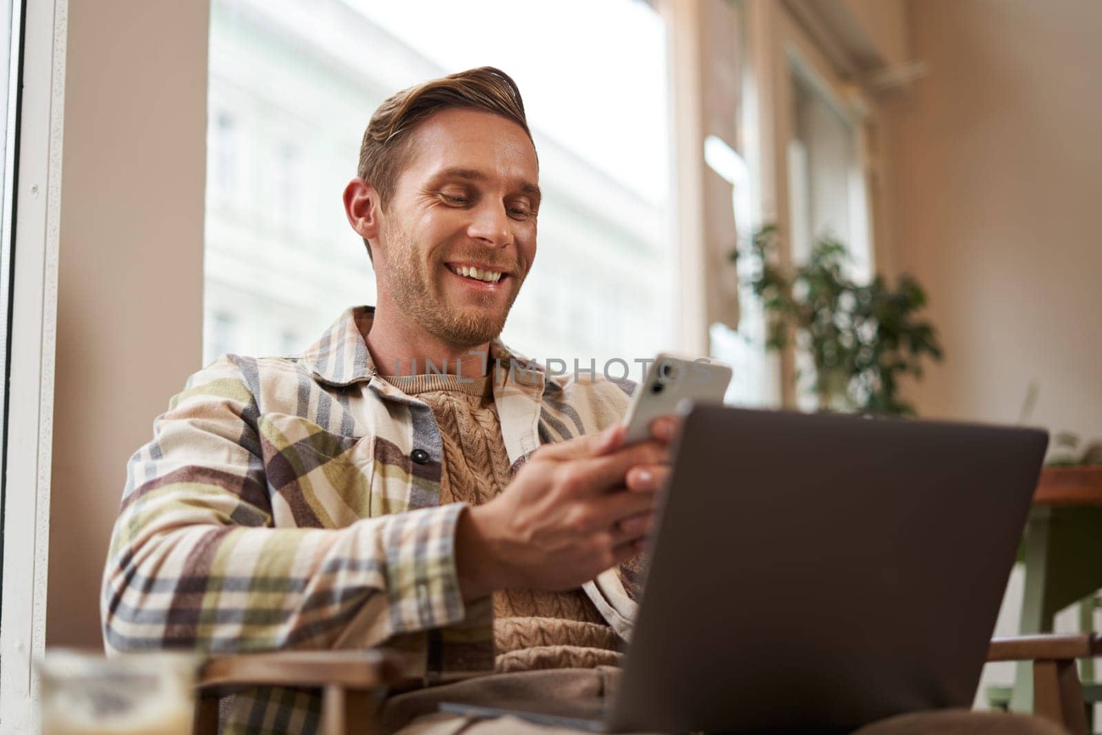 Close up portrait of handsome smiling man with laptop, cafe visitor sitting in chair, using smartphone, looking happy, messaging with friends while enjoying cup of coffee.