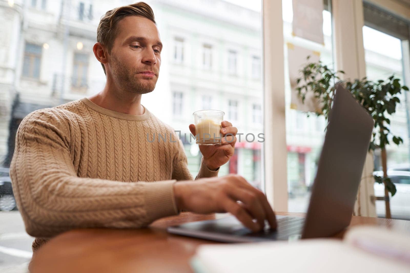Portrait of handsome young man sitting in cafe with laptop. Digital nomad, freelancer working outdoors from coffee shop, typing something by Benzoix