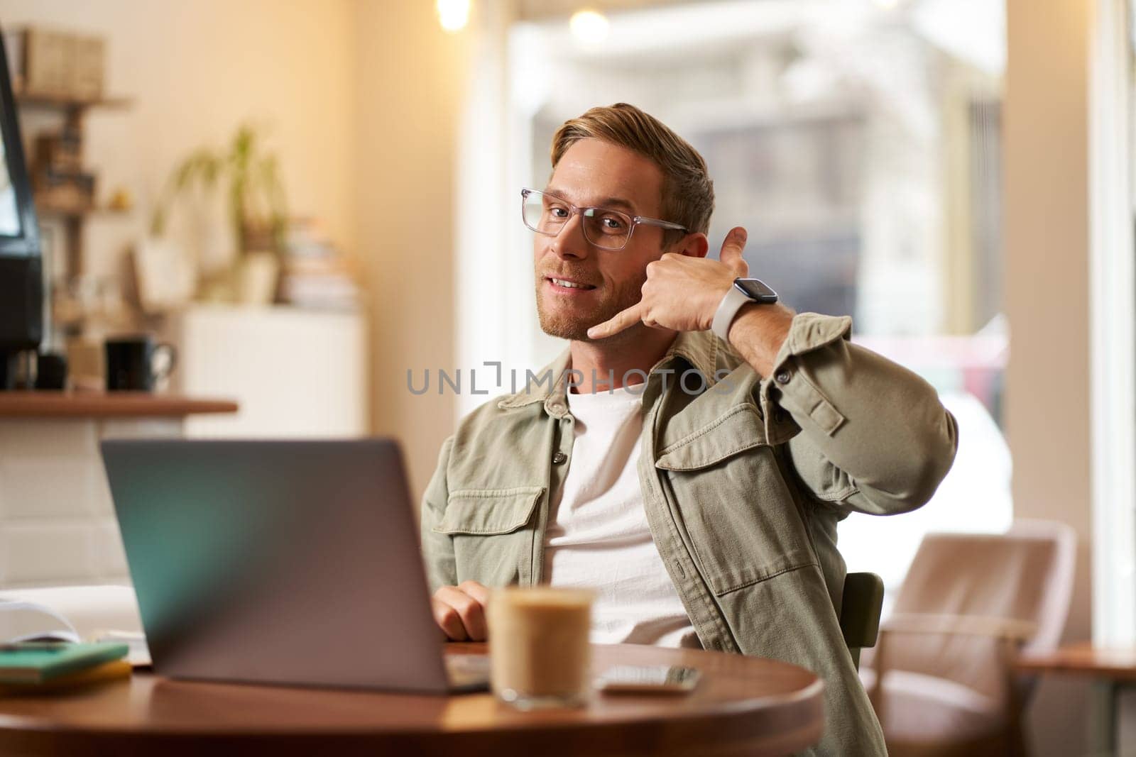 Portrait of young handsome man in glasses, businessman working in cafe, sitting with laptop, showing phone call hand gesture, advertising his service by Benzoix