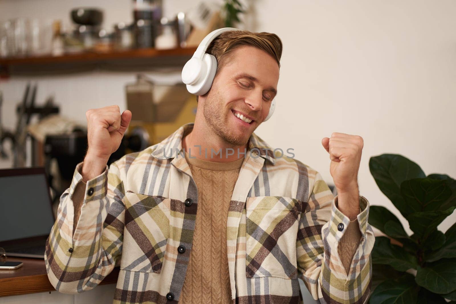 Portrait of handsome happy guy, freelancer working and listening to music in wireless headphones, dancing on his chair in cafe, enjoying favourite song in earphones.