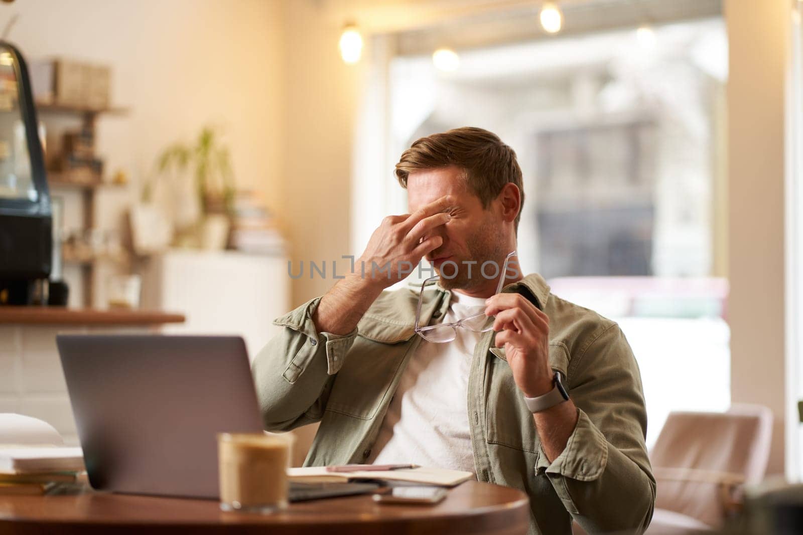 Portrait of tired man rubbing his eyes after looking at laptop screen and working on computer, sitting in cafe, having a break, taking off his glasses.