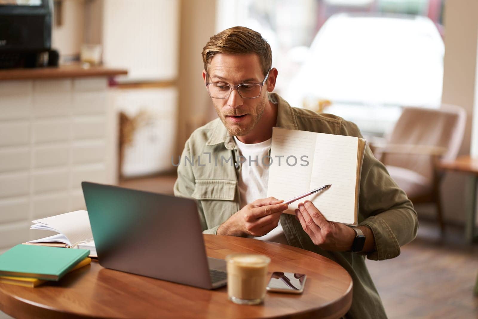 Portrait of young handsome man in glasses, private tutor teaching student online, pointing at his notebook, showing something, video chats via laptop application, working remotely in cafe.