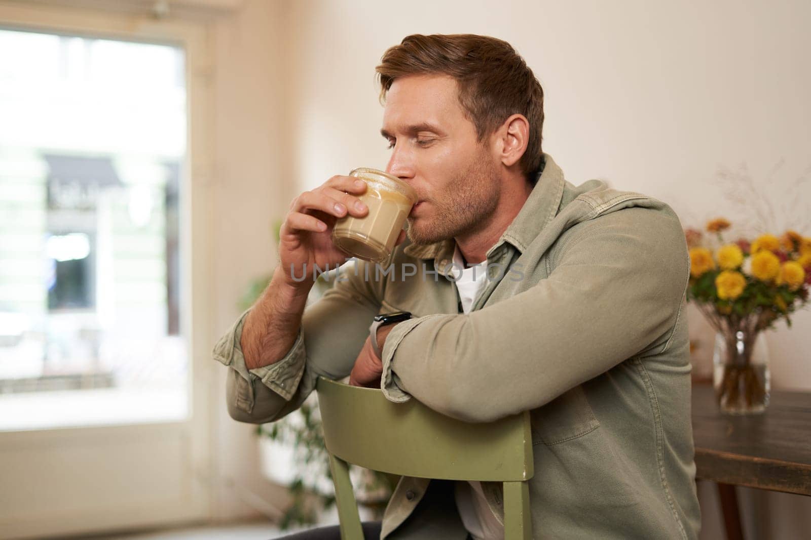 Portrait of handsome blond man in cafe, sits on chair, enjoys the taste of delicious freshly brewed cup of coffee.