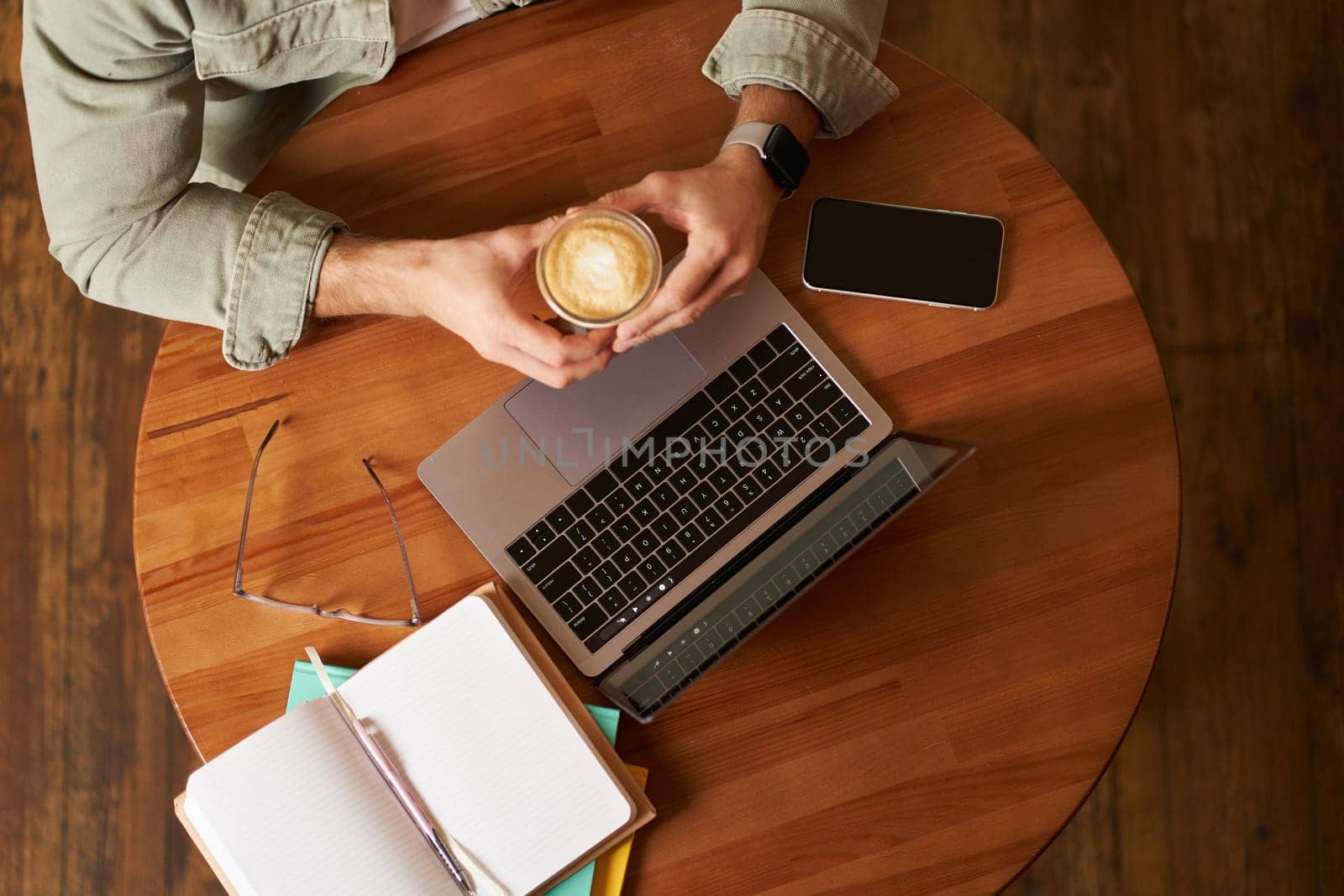Top view. Male hands holding cup of coffee, man sitting at round table in a cafe, working on laptop, had his smartphone and notebook, drinking cappuccino by Benzoix