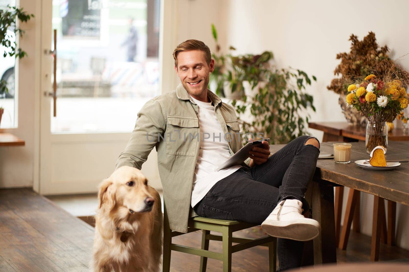 Portrait of happy smiling young man, cafe visitor, sitting in coffee shop with his dog, petting golden retriever, holding digital tablet, reading news or browsing on internet by Benzoix