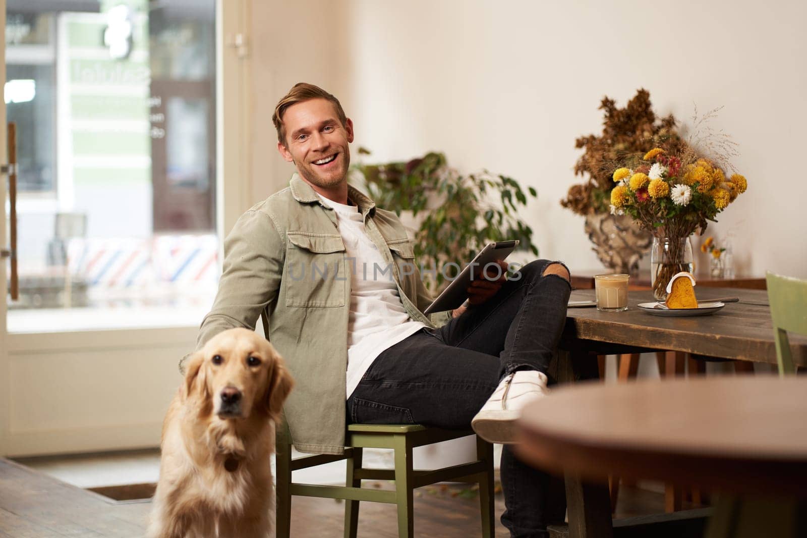 Portrait of handsome young man, cafe visitor, sitting with his dog, petting golden retriever and reading news on digital tablet, concept of lifestyle and pet-friendly public places.