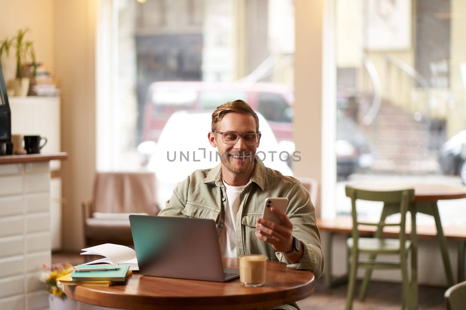 Portrait of handsome man in glasses, sits in cafe with laptop, smiles while reads a message on phone, checks his smartphone app, works from coffee shop.