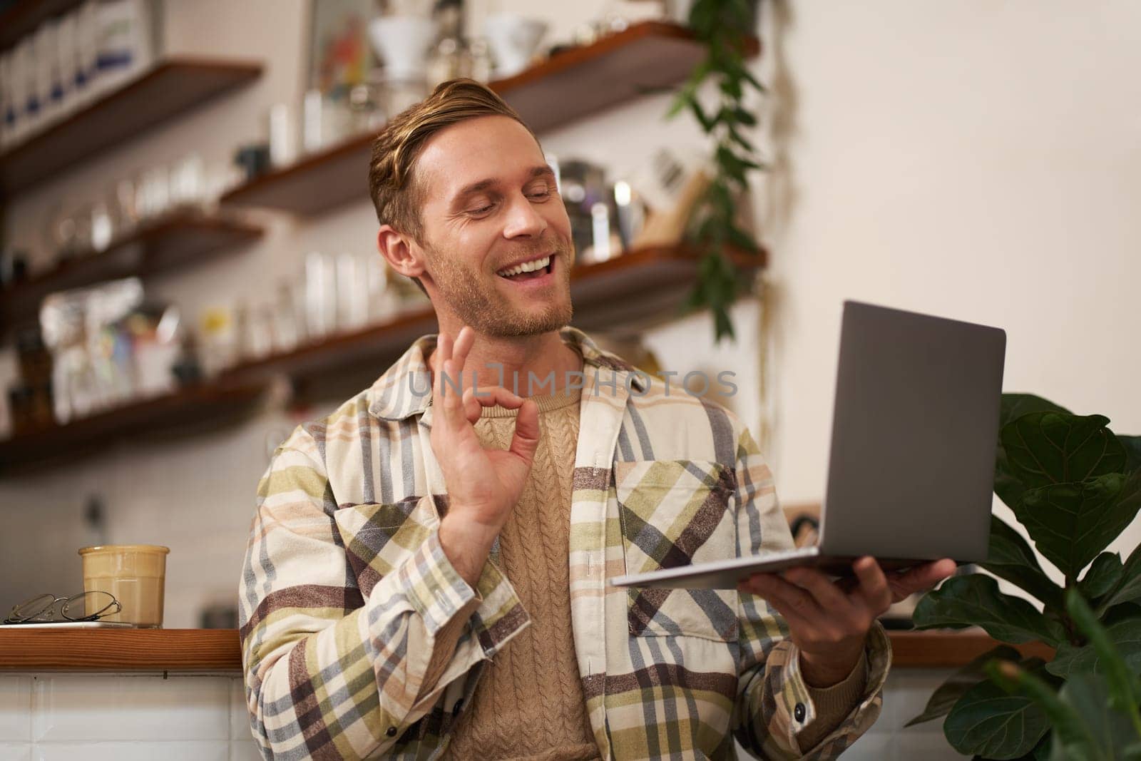 Portrait of young smiling man, coworker joins online meeting from cafe, video chats on laptop, shows okay, ok hand sign in approval, drinking coffee and working remotely by Benzoix