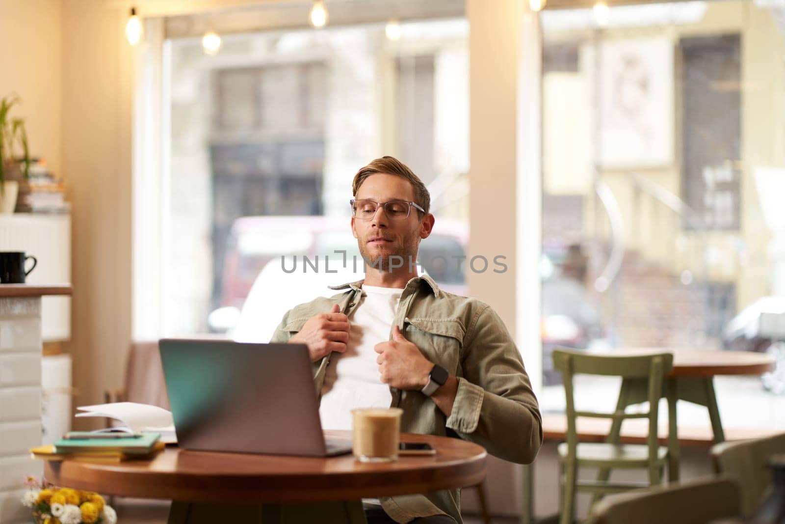 Portrait of businessman in glasses, sits in cafe with laptop, looks concentrated at screen with his project task, works remotely from coffee shop.