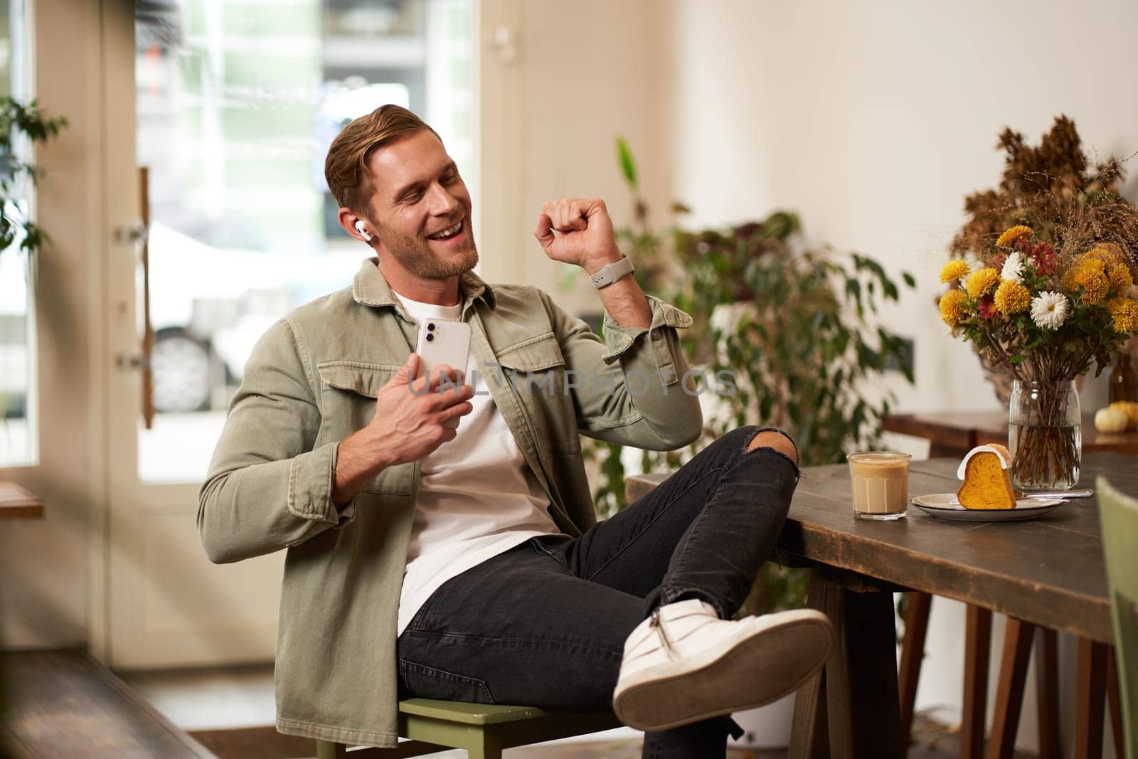 Portrait of handsome happy man in cafe, listens to music in wireless earphones, holding smartphone, connects to public wifi and enjoys favourite song, relaxing in coffee shop.