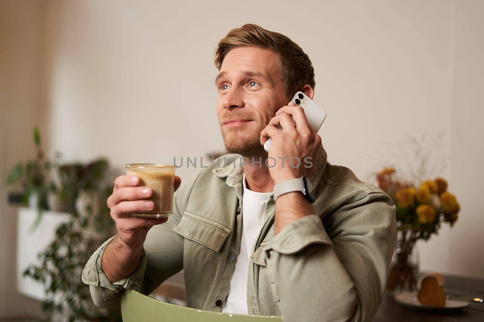Image of smiling, good-looking young blond man with phone, drinking coffee in cafe, talking to someone over the telephone, answering a call.