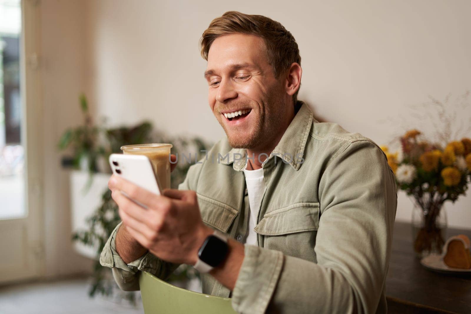 Image of handsome young man in cafe, looking at his mobile phone, using smartphone app, drinking coffee.