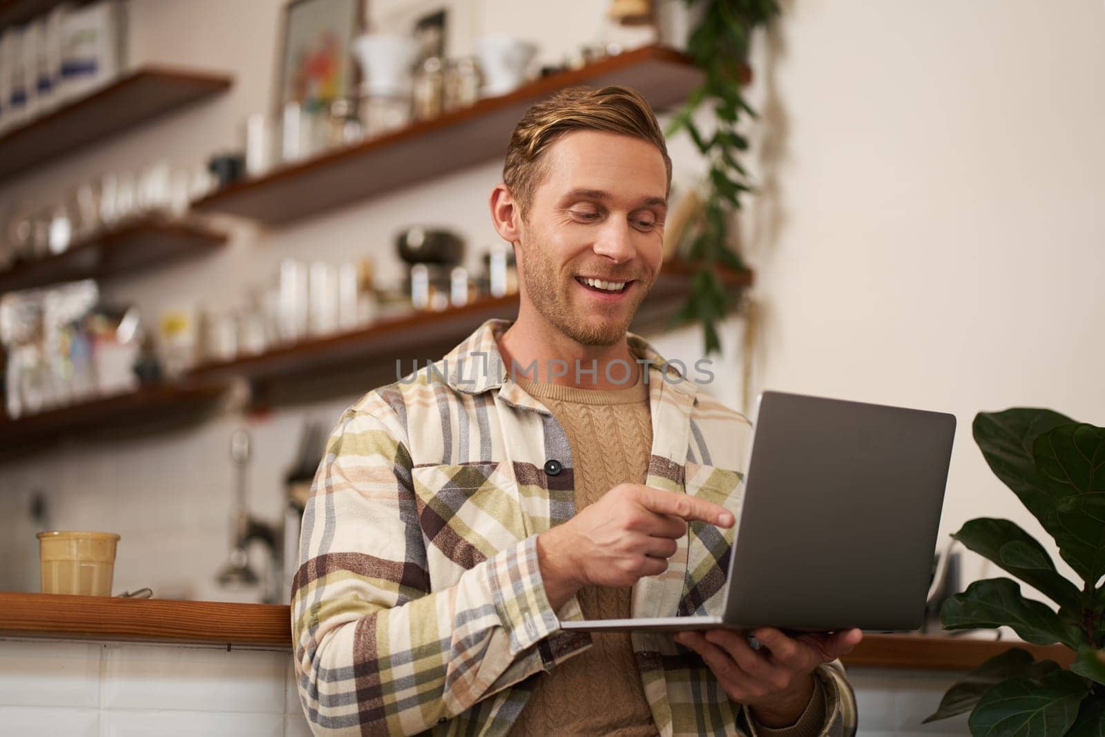 Image of young man in coffee shop, connects to video call, talking online, chats with friends via laptop and smiles.