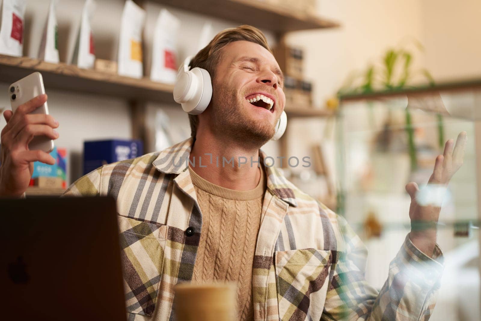 Portrait of handsome happy guy, freelancer working and listening to music in wireless headphones, dancing on his chair in cafe, enjoying favourite song in earphones.