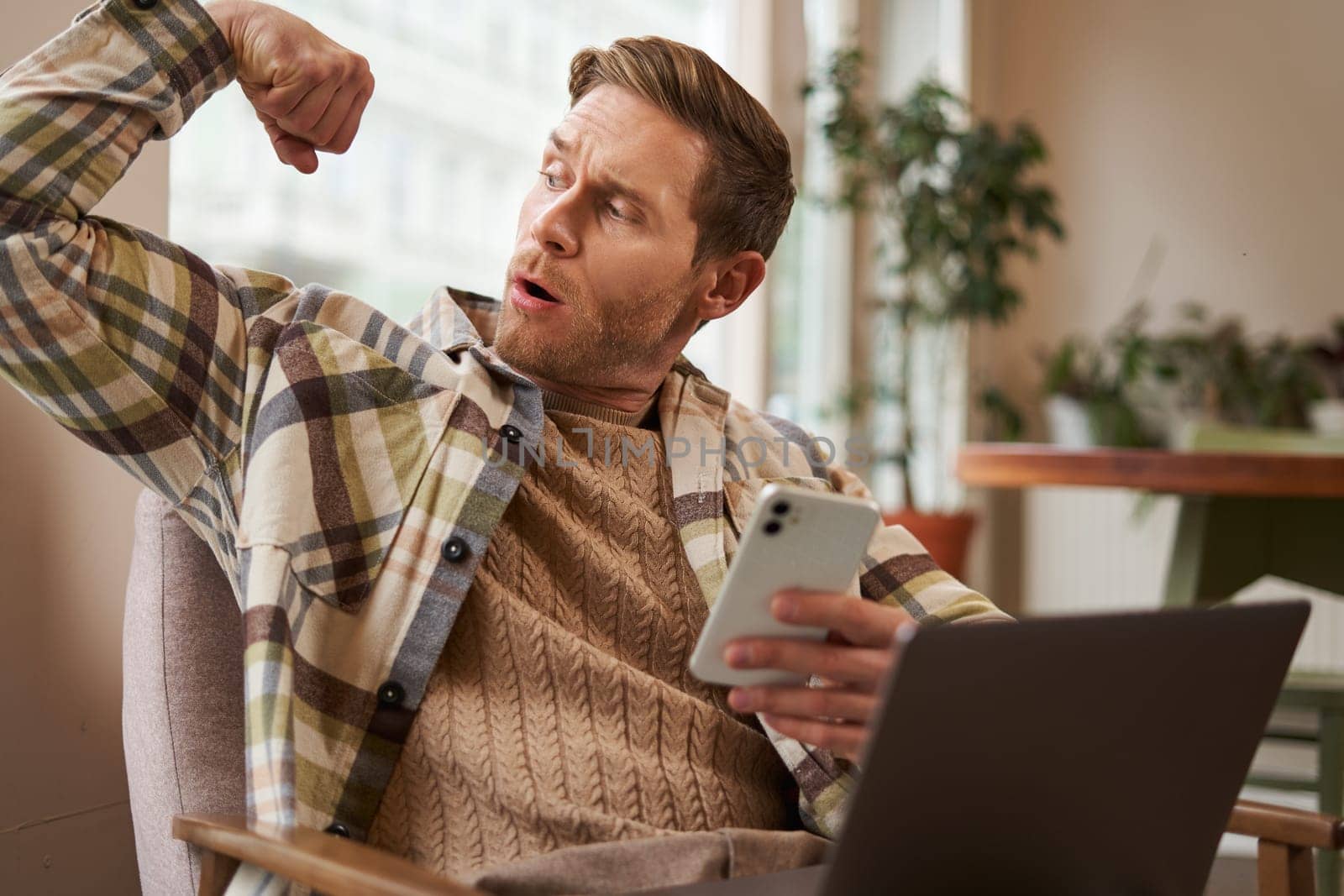 Portrait of young athletic guy sitting in cafe with laptop and smartphone, showing his biceps, flexing muscles and looking pleased at himself.