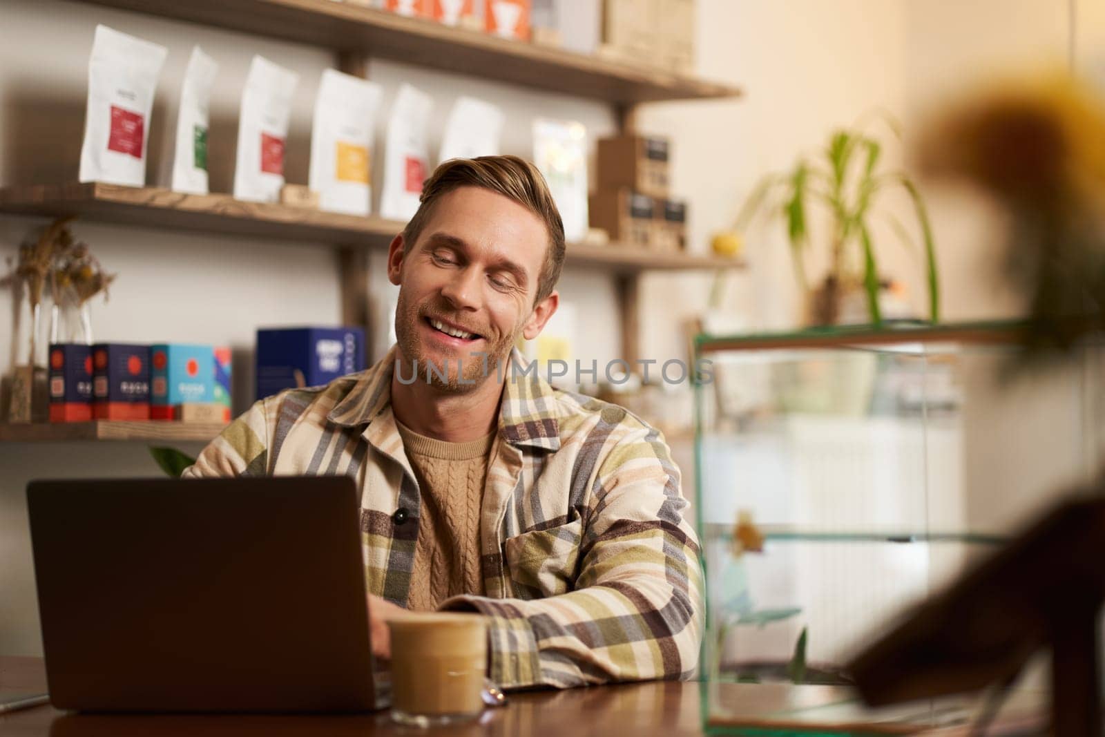 Portrait of handsome young digital nomad, man working in cafe on laptop, looking happy and pleased with his online project, sitting in co-working coffee shop by Benzoix