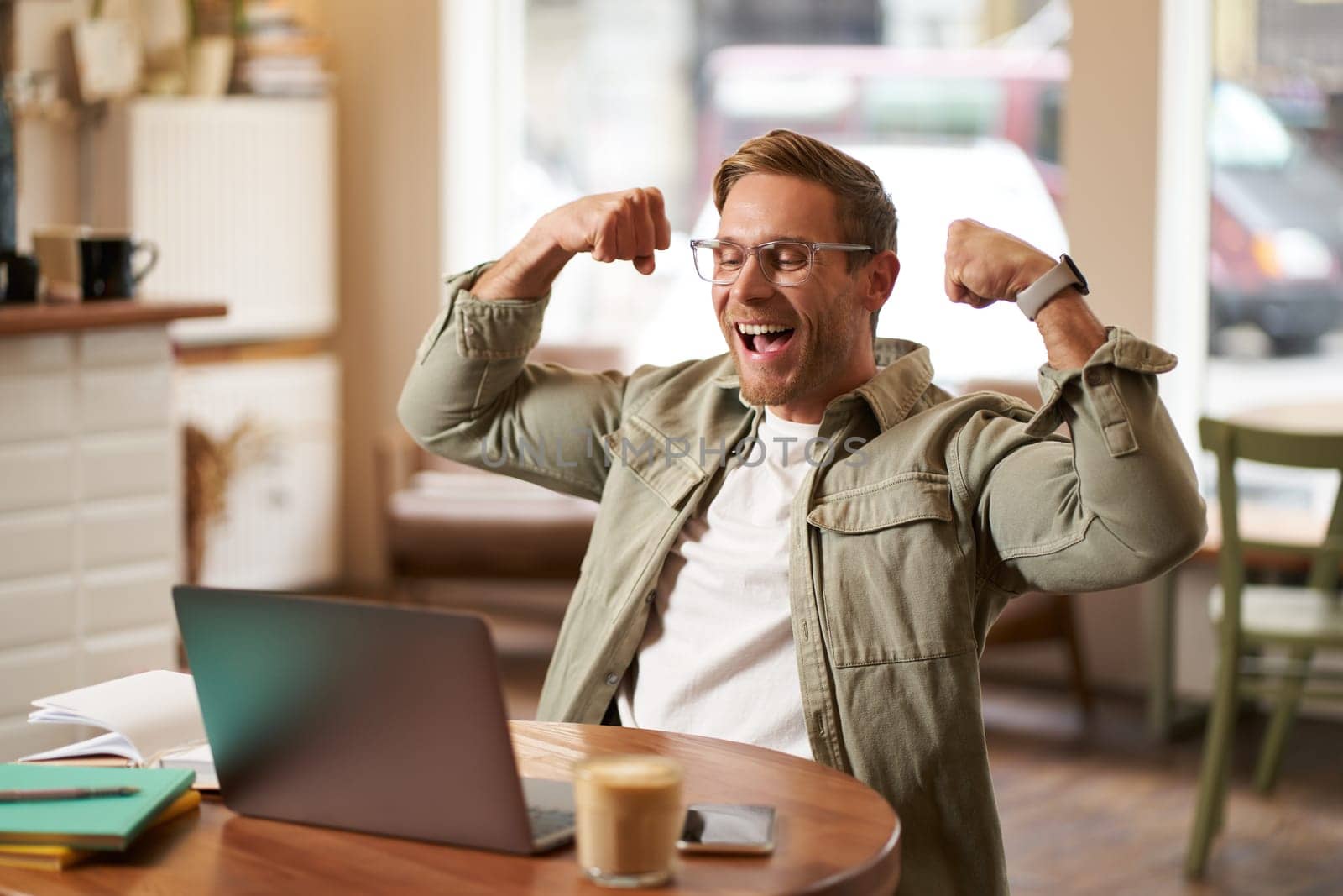 Portrait of happy young man in glasses rejoicing, sitting in cafe, watching sports game on laptop, celebrating, showing big muscles and laughing.