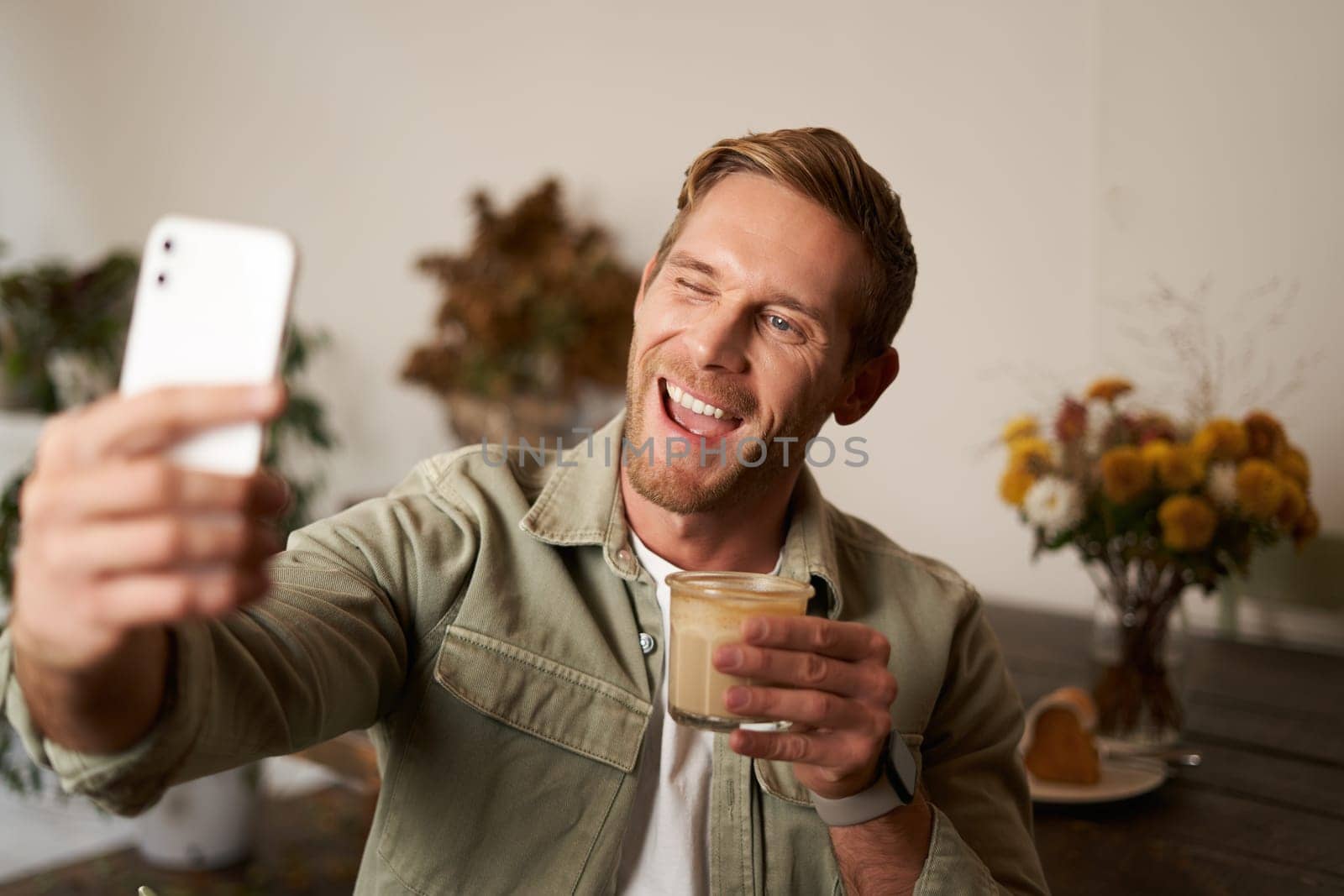 Close up portrait of happy, charismatic young blond man, taking selfie on smartphone, posing with cup of coffee, enjoys his drink in cafe, recommending a place to his followers online by Benzoix