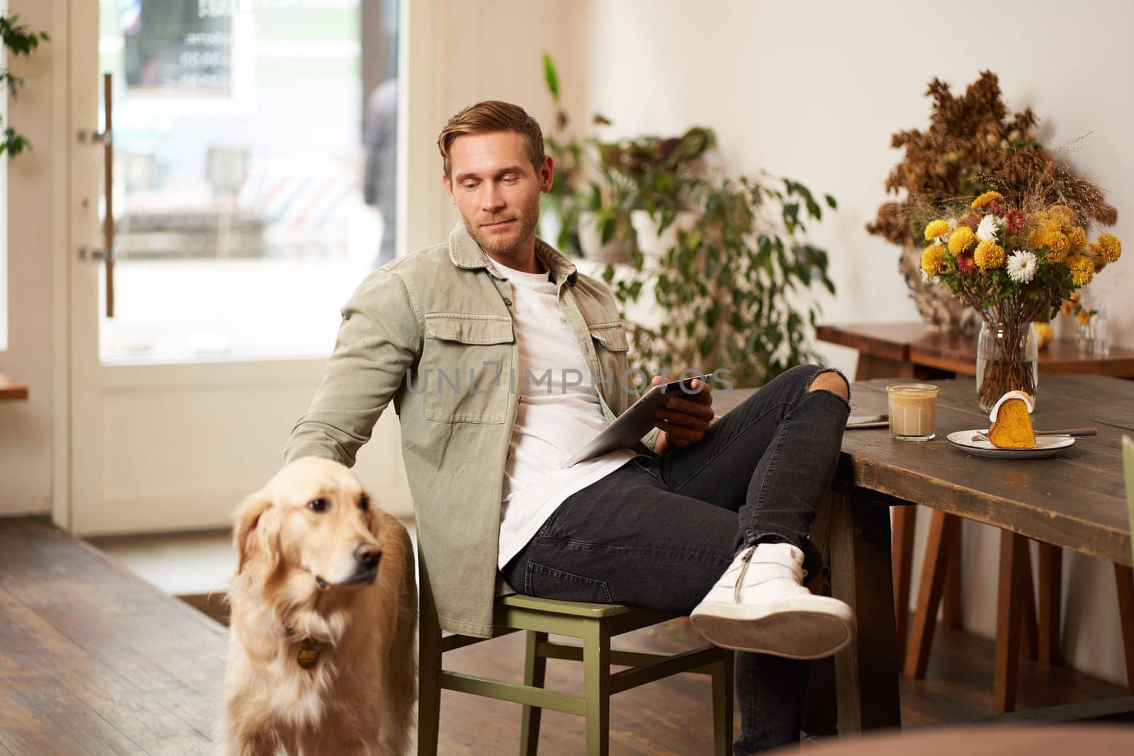 Portrait of handsome young man sits in cafe with his dog, petting golden retriever, reading news on tablet, relaxing with cup of coffee.