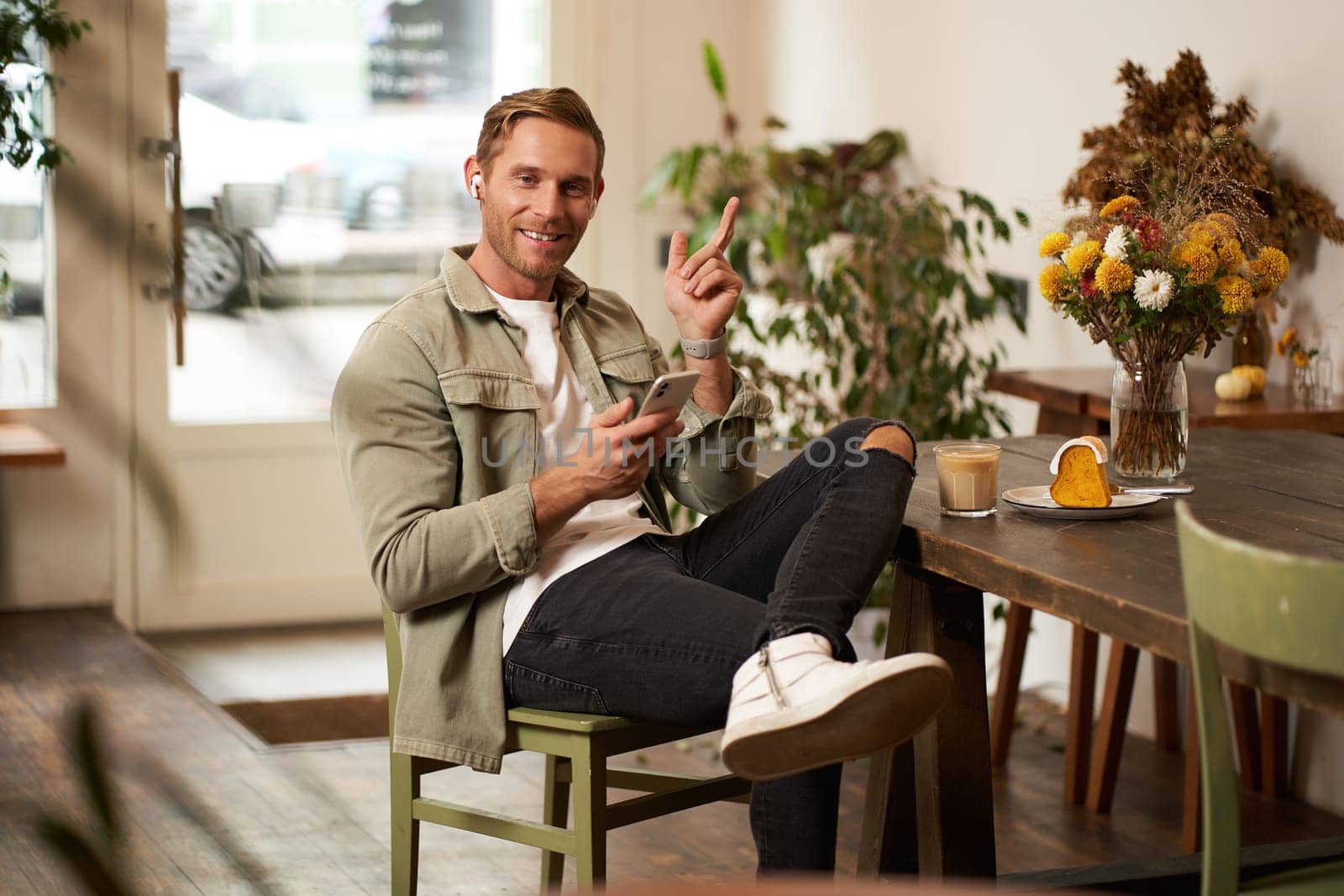 Portrait of happy handsome young man, sitting in cafe with mobile phone, wearing wireless headphones, listening to music or podcast while relaxing in coffee shop.