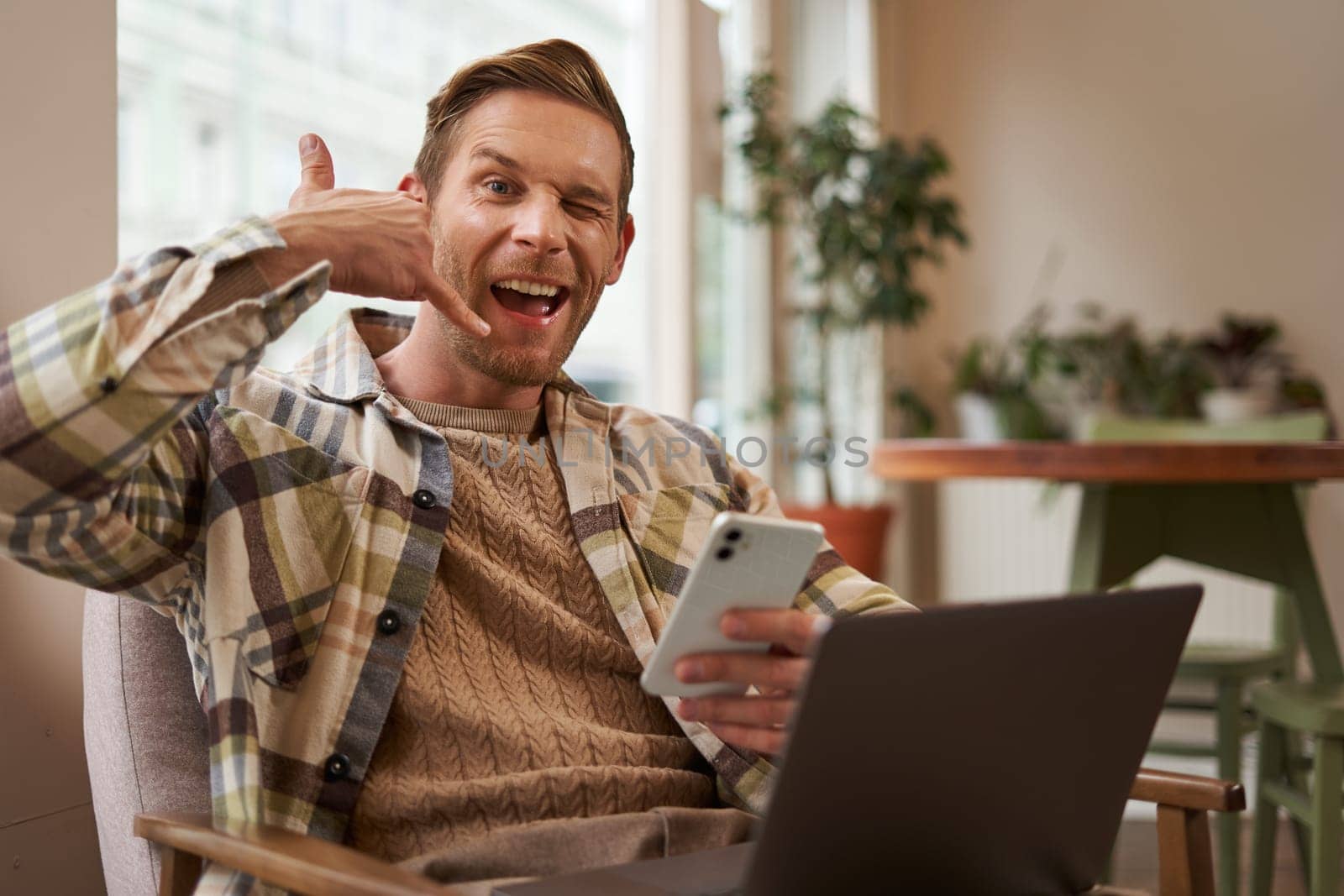 Portrait of handsome male cafe visitor, businessman working on laptop, holding smartphone, showing mobile phone hand sign, give me a call gesture.