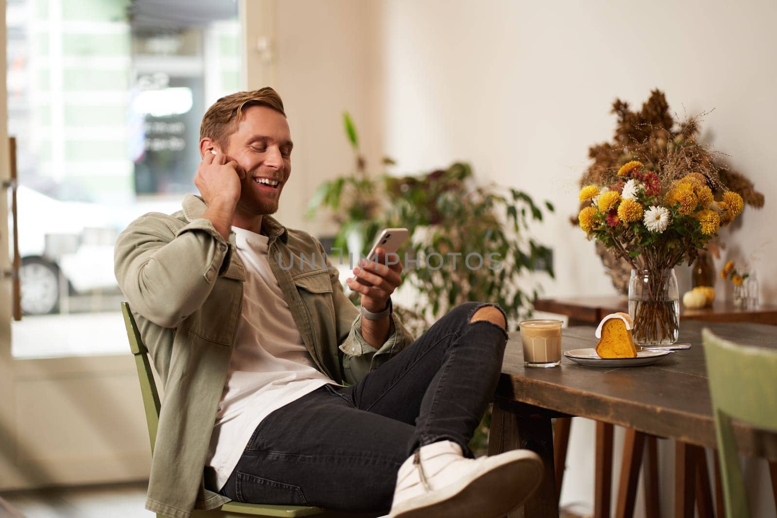 Lifestyle shot of handsome young man, looking at his mobile phone, listening to music in wireless earphones, sitting in a cafe, drinking coffee.