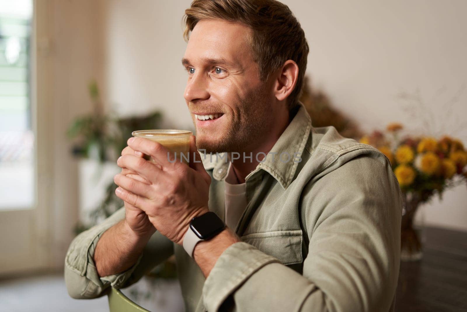 Leisure, people and lifestyle concept. Happy handsome young man in cafe, sitting on chair and smiling, holding glass of coffee, looking aside, relaxing with cup of cappuccino.