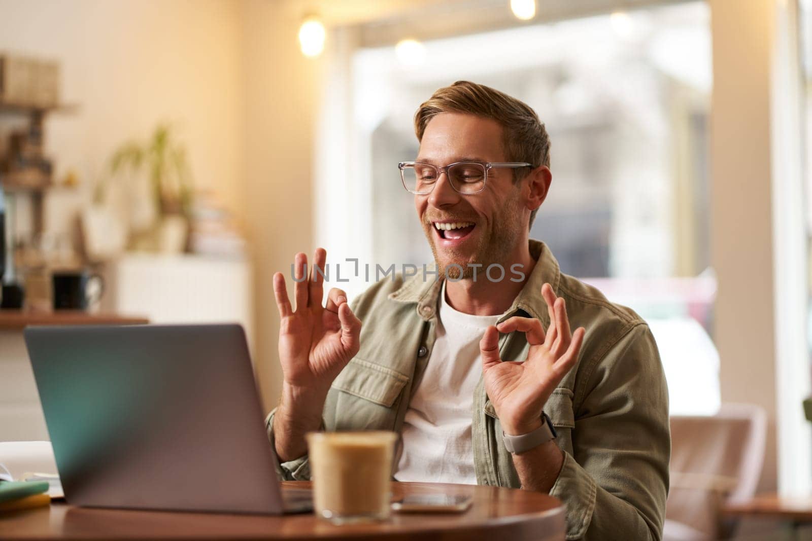 Portrait of young freelancer, digital nomad having an online meeting, video chats, shows okay sign at laptop, confirm something, makes ok hand gesture, sits in cafe, works on remote.