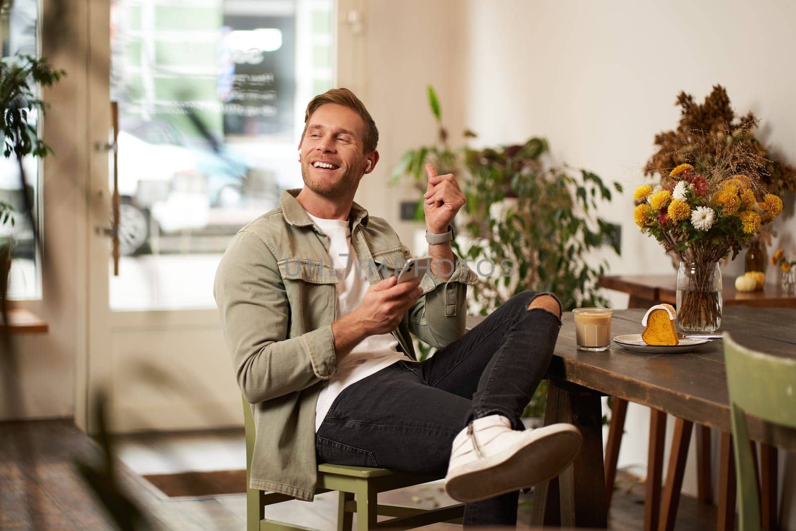 Portrait of young handsome man in casual clothes, sits in cafe and enjoys favourite song, listens to music in wireless headphones, vibing to the relaxing tunes in earphones, holding smartphone.