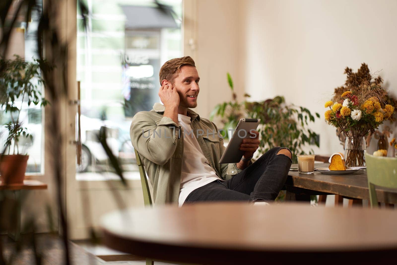 Portrait of handsome smiling young man, sitting in coffee shop, using digital tablet, video chats with someone from a cafe, wearing wireless headphones by Benzoix