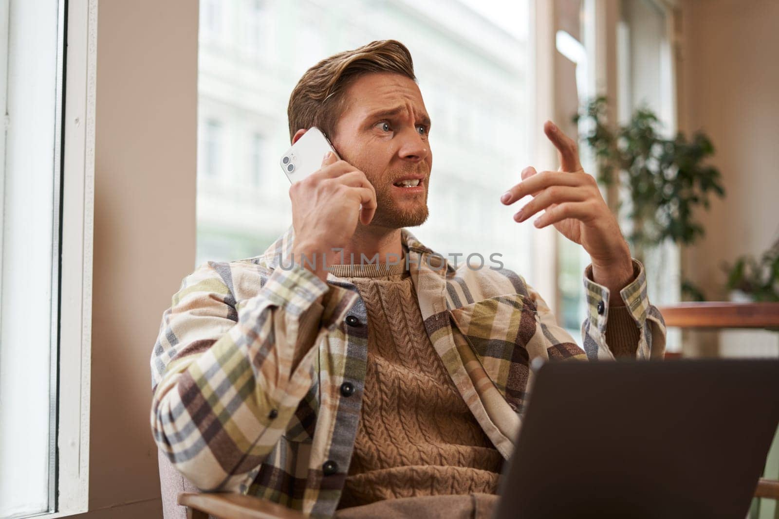 Image of man feeling stressed while talking over the phone. Businessman sitting in cafe with laptop, answering a call, hear bad news over the telephone.
