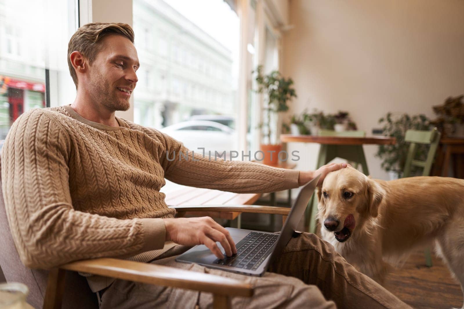 Handsome young man working in cafe with a dog, sitting on chair and using laptop, petting his golden retriever in animal-friendly co-working space by Benzoix