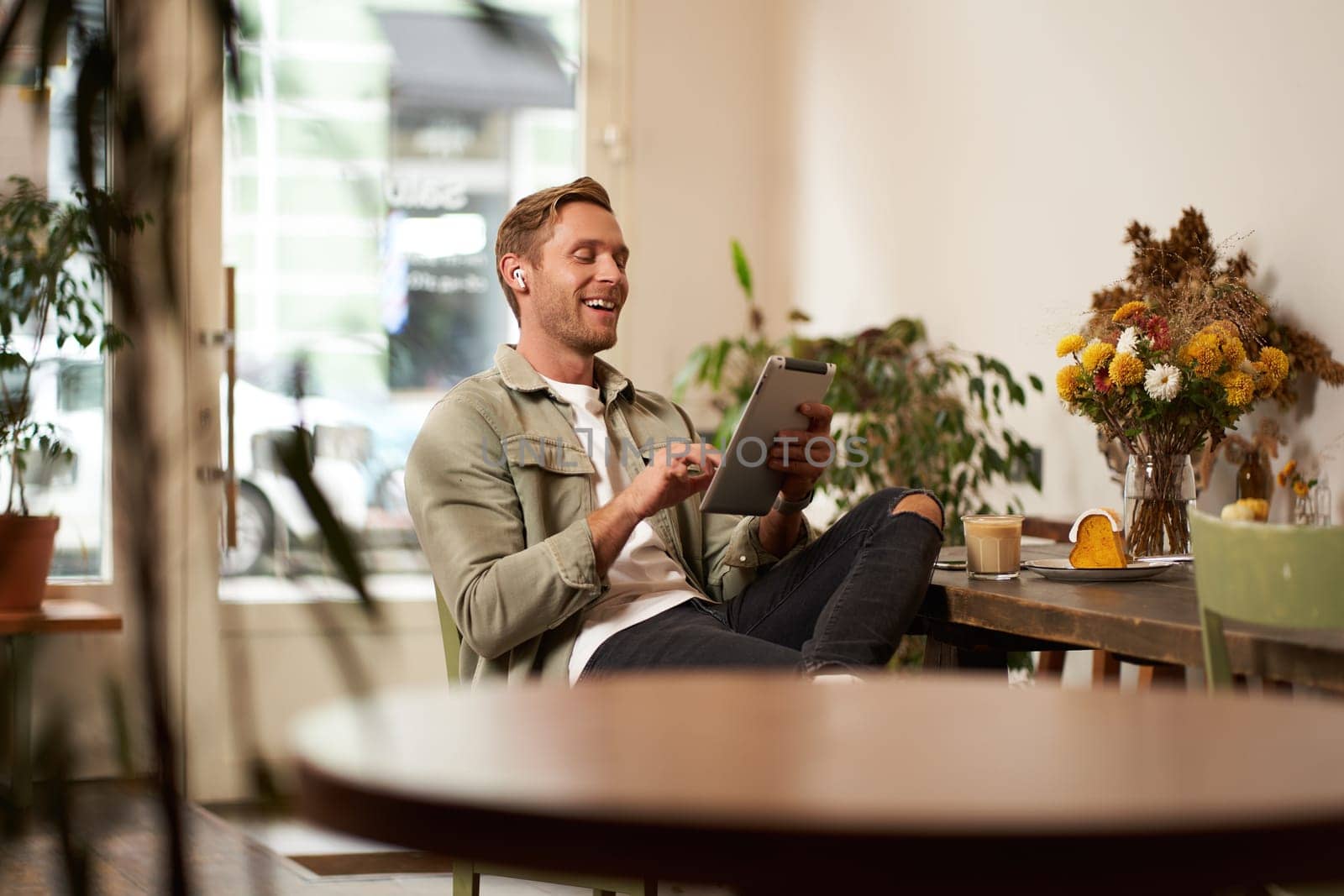 Portrait of handsome happy guy, young man sits in cafe, watching videos on digital tablet, wearing wireless headphones, laughing and smiling, spending time in coffee shop by Benzoix