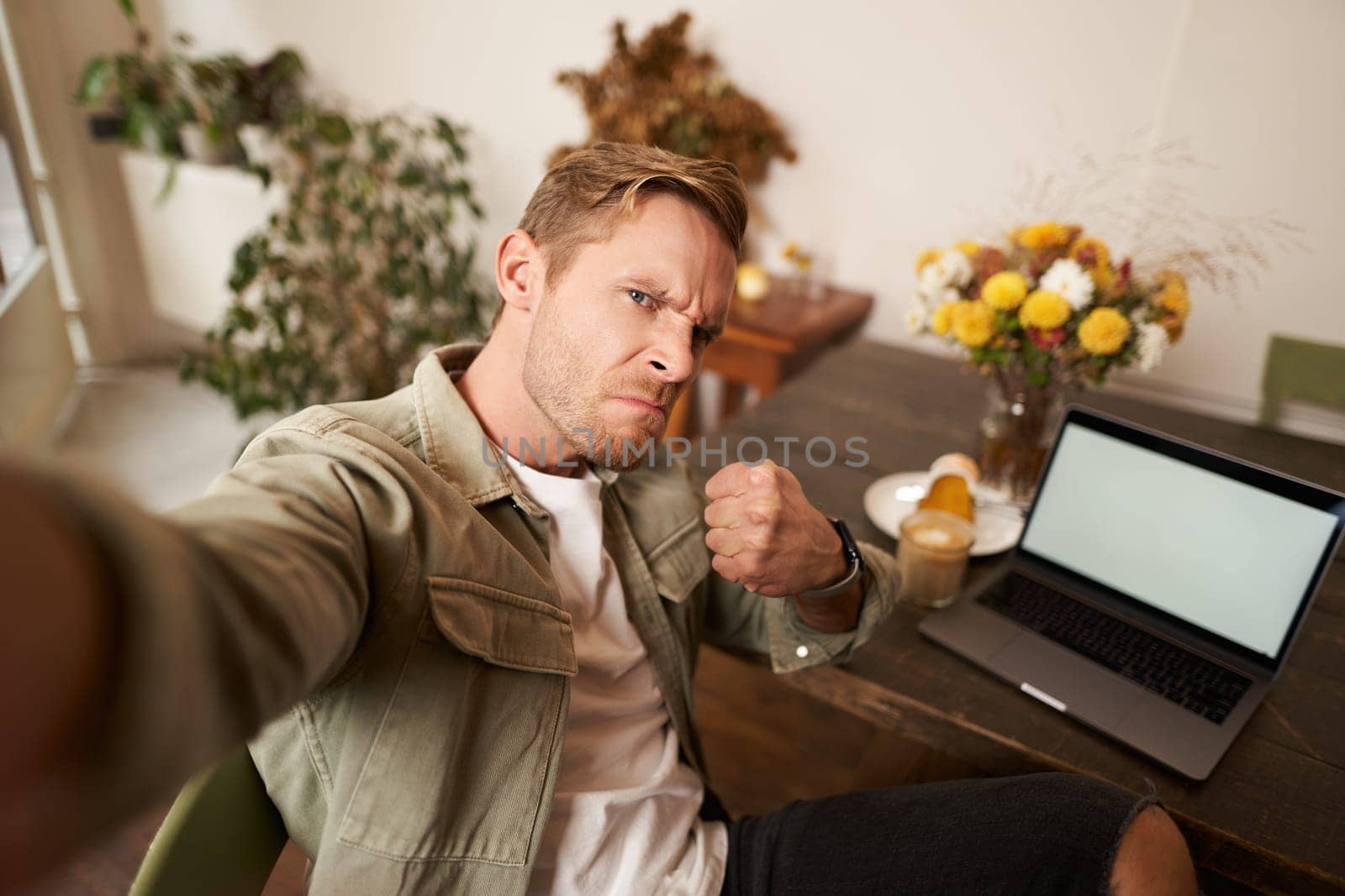 Portrait of handsome man making angry threatening face for selfie photo, taking picture on smartphone app in cafe, sitting near laptop.